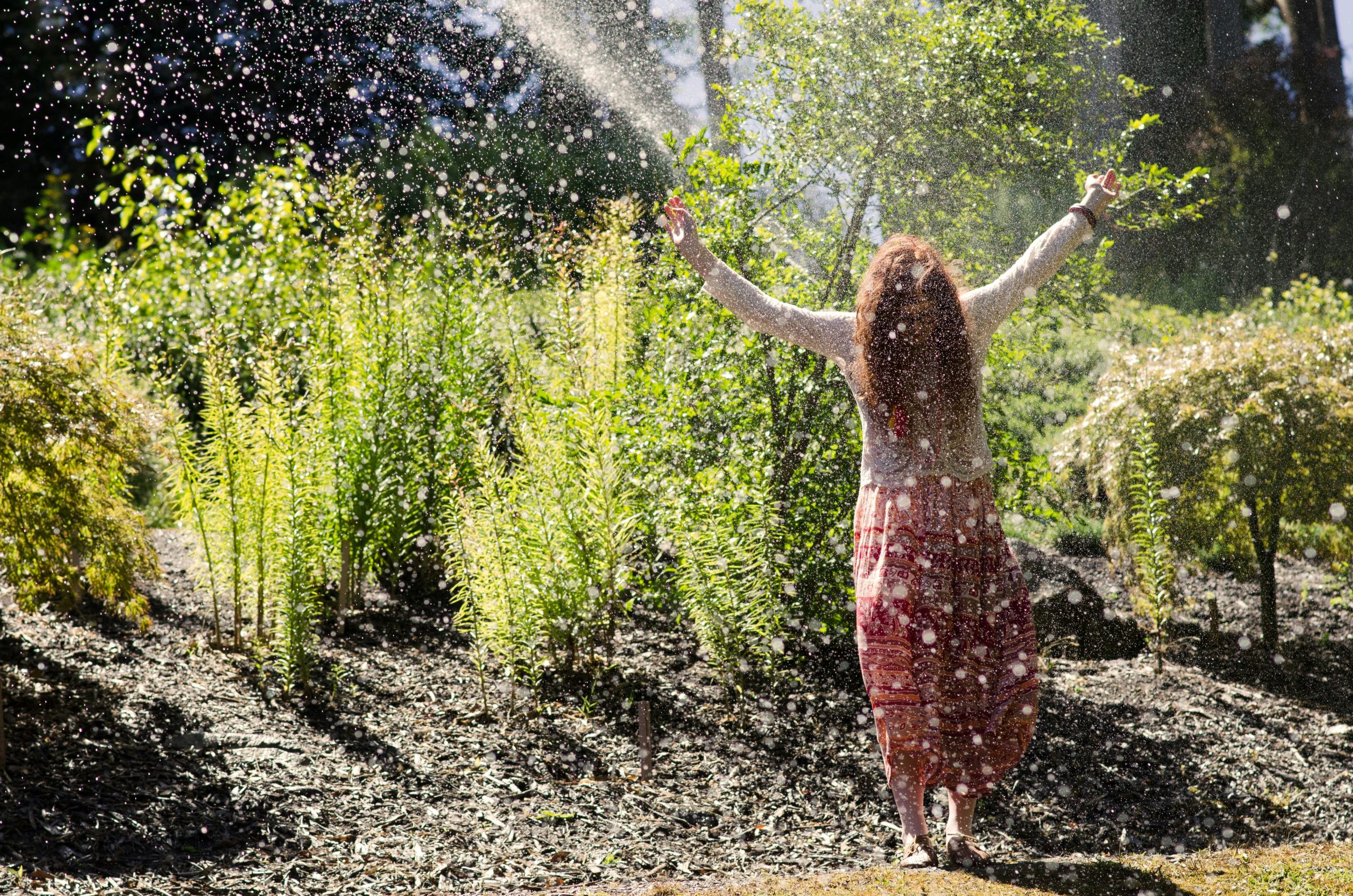 Woman enjoying outdoor nature therapy with arms raised under a water sprinkler, symbolizing freedom and healing in addiction recovery
